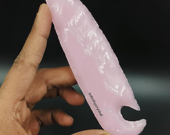 May include: A pink, translucent, arrowhead-shaped crystal with a hole at the bottom. The crystal is being held in a hand. The text "Infinitycrystal" is visible on the crystal.