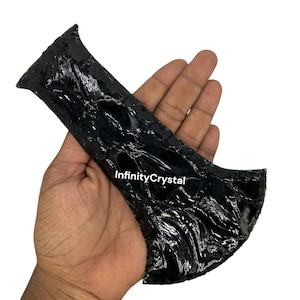 May include: A polished black obsidian arrowhead with a textured surface. The arrowhead has a unique shape with a flared top and a pointed bottom. The text "InfinityCrystal" is visible on the surface.