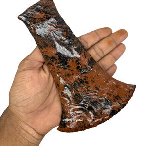 May include: A brown and black stone axe head with a textured surface. The axe head is held in a hand against a white background. The text "InfinityCrystal" is visible on the axe head.