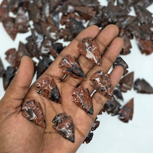May include: A collection of red and black obsidian arrowhead replicas. Several arrowheads are held in a hand, with many more scattered in the background. The arrowheads have a glossy finish and are made of a stone material.
