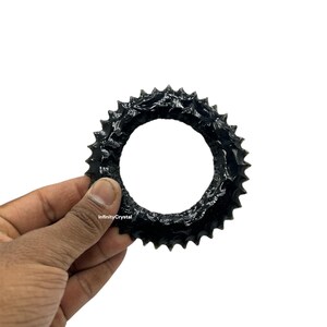 May include: A black, circular object with a spiky, textured edge is held against a white background. The object has a large, open center. The text "InfinityCrystal" is visible on the object.