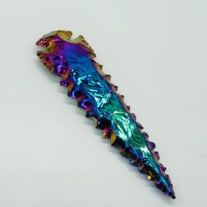 May include: A dichroic glass arrowhead with a vibrant, iridescent finish. The arrowhead is a mix of blues, greens, purples, and golds. The edges are jagged and textured, and the tip is pointed.