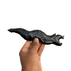 May include: A black obsidian dragon carving. The dragon is held in a hand against a white background. The carving has a textured surface and a detailed dragon head. The text "InfinityCrystal" is visible on the carving.