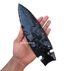 May include: A large, polished black obsidian arrowhead with a sharp point and notched base. The arrowhead is held in a hand against a white background. The text "INFINITYCRYSTAL" is visible on the arrowhead.