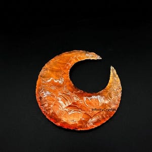 May include: An orange crescent-shaped crystal with a textured surface. The crystal is set against a black background. The text "InfinityCrystal" is visible on the right side of the crystal.