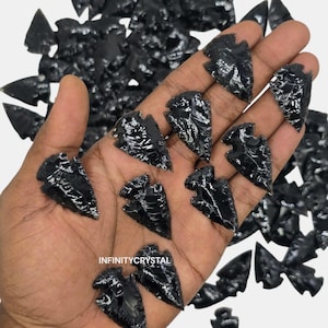 May include: A collection of polished black obsidian arrowheads. The arrowheads are held in a hand, with many more scattered in the background. The arrowheads have a glossy, reflective surface. The text "INFINITYCRYSTAL" is visible.