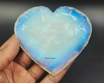 May include: A heart-shaped piece of iridescent opal with a blue and white color scheme. The opal is smooth and polished, and it has a natural, organic shape. The opal is being held in a hand, and the background is a dark, blurry surface.