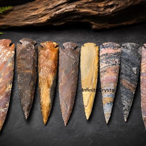 May include: Eight hand-knapped stone arrowheads arranged in a row, featuring varied earth tones including tan, brown, grey, and banded patterns. Each arrowhead has a pointed tip and a notched base. The text InfinityCrystal is visible on one.