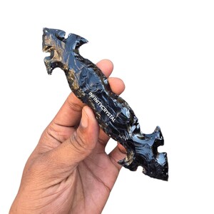 May include: A black obsidian arrowhead with a textured surface and intricate carved details. The arrowhead is held in a hand and features the text "INFINITYCRYSTAL".
