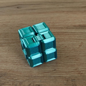 Op de afbeelding: Een turquoise fidget cube ligt op een houten oppervlak. De kubus bestaat uit acht verbonden blokken, elk rechthoekig. De blokken hebben een textuur en zijn ontworpen voor tactiele manipulatie en stressverlichting.