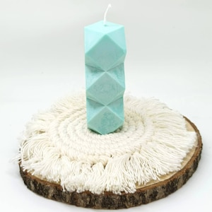 Könnte beinhalten: Eine hellblaue geometrische Kerze auf einem weißen Makramee-Untersetzer mit Holzbasis.