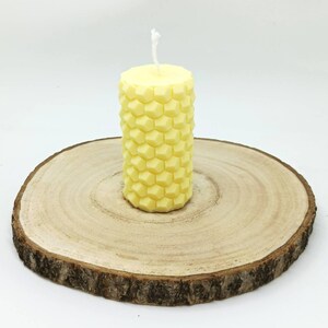 Könnte beinhalten: Eine gelbe, zylindrische Kerze mit einem Wabenmuster. Die Kerze steht auf einer runden Holzscheibe mit sichtbaren Jahresringen. Ein weißer Docht ragt aus der Oberseite heraus. Der Hintergrund ist weiß.