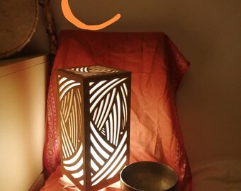 以下が含まれることがあります： 幾何学模様の入った木製のランプが点灯し、温かい光を放っています。ランプは赤い布の表面に置かれ、歌碗と木製の槌の隣にあります。
