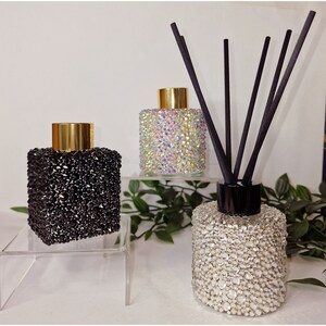 Könnte beinhalten: Drei Glasdiffuserflaschen mit goldenen Deckeln und schwarzen Stäbchen. Die Flaschen sind mit schwarzen, schillernden und klaren Strasssteinen bedeckt.