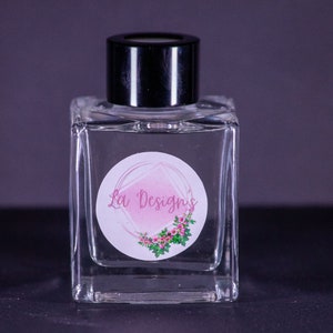 May include: A clear glass bottle with a black lid and a white label with a pink watercolor design and the text "La' Designs".