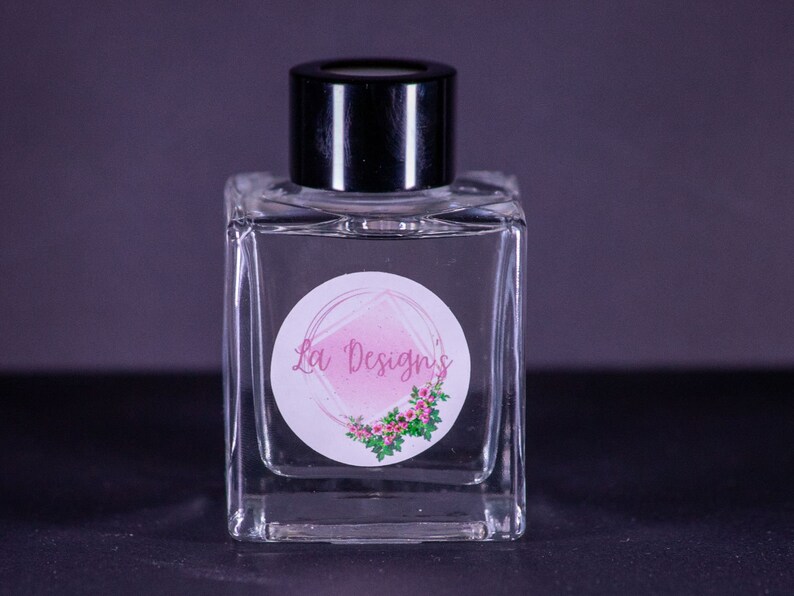 May include: A clear glass bottle with a black lid and a white label with a pink watercolor design and the text "La' Designs".