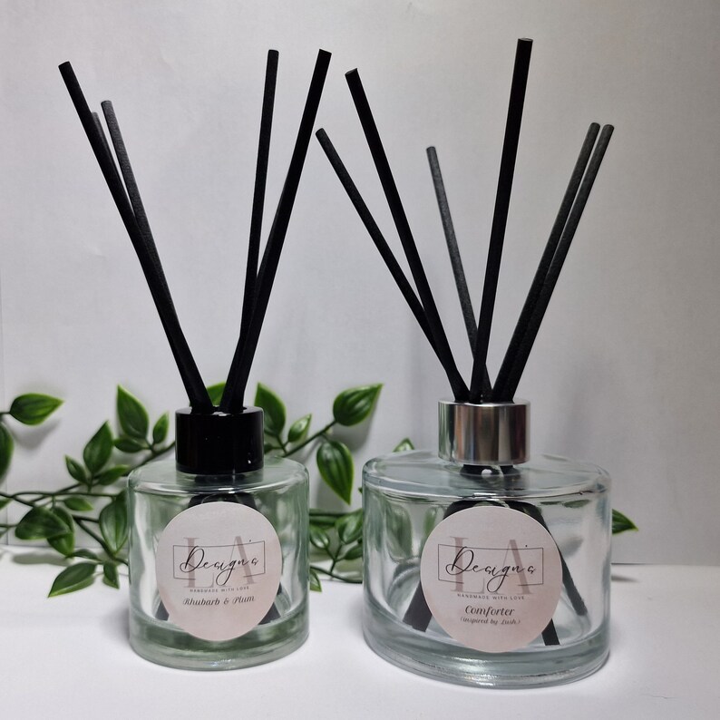 May include: Two clear glass bottles with black diffuser sticks and labels. The labels are white with black text and a pink circle. The left bottle says "Rhubarb & Plum" and the right bottle says "Comforter".