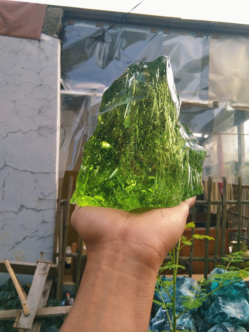 May include: A large, green, glass rock with a rough, textured surface. The rock is being held in a person's hand.