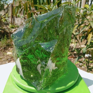 May include: A large, green, translucent piece of glass on a green scale. The glass has a rough, irregular shape and appears to be naturally formed.