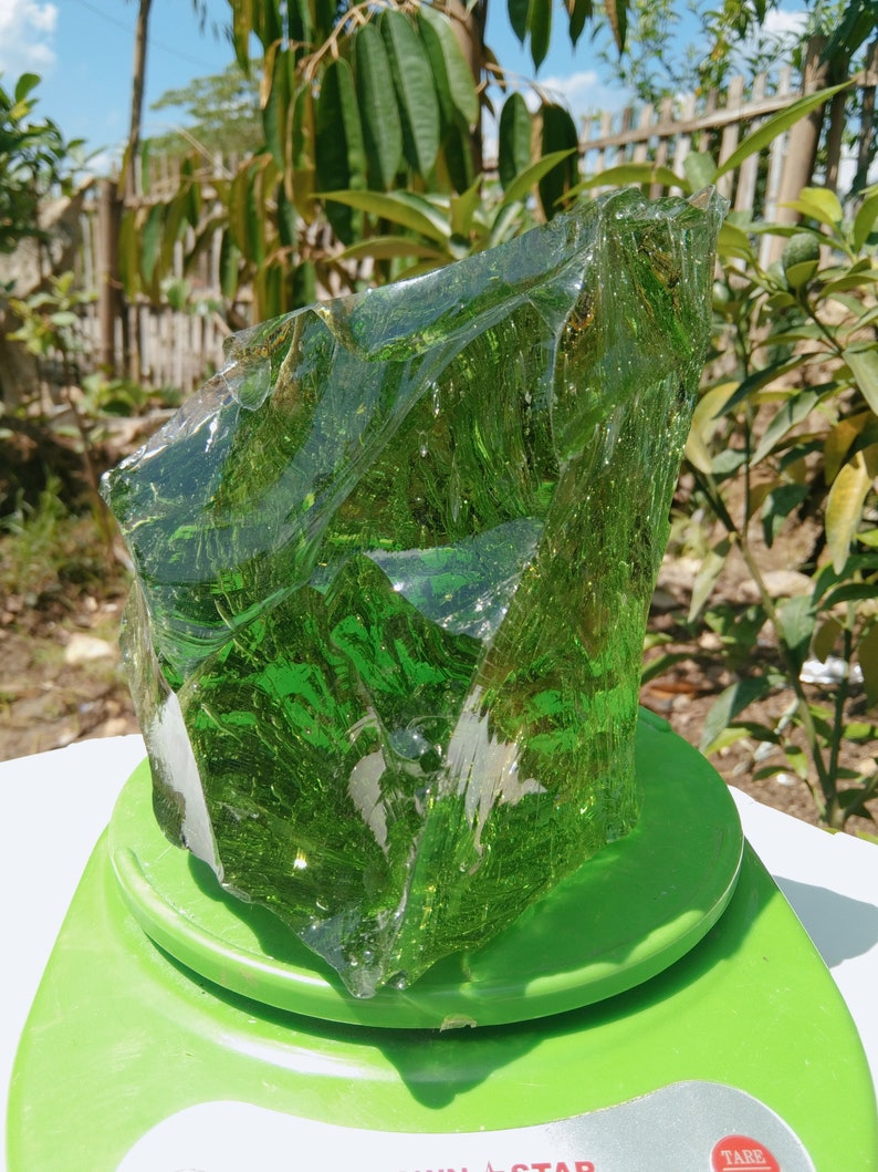 May include: A large, green, translucent piece of glass on a green scale. The glass has a rough, irregular shape and appears to be naturally formed.