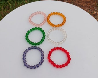May include: Six colorful beaded bracelets on a white surface. The bracelets are made of glass beads in pink, orange, green, clear, purple, and red.