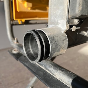 May include: Close-up of a black plastic dust port on a power tool. The port is round and has a lip around the edge. The port is attached to a metal pipe.