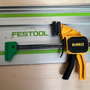 May include: A yellow and black spring clamp with a black handle and the word "DEWALT" printed on the side. The clamp is attached to a silver and green track with the word "FESTTOOL" printed on it.