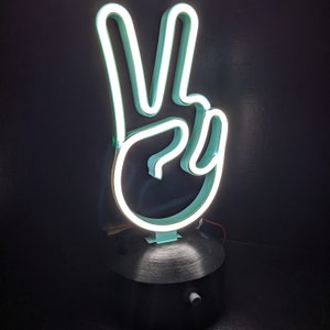 May include: A white neon peace sign on a black base. The neon sign is lit up and the base has a switch on the bottom.