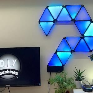 May include: A wall with a blue and black triangular light fixture, a black computer monitor with the text "D.I.Y. machines" on the screen, a small potted plant, and a white potted bonsai tree.