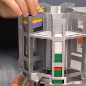 May include: A 3D printed, grey, multi-level, rotating structure with colourful blocks inserted into the slots. The structure is part of a DIY machine.