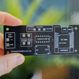 Könnte beinhalten: Eine schwarze Leiterplatte mit weißem Text und Komponenten, darunter "WS2811", "LED-DATA" und "D1 mini". Sie hat verschiedene Ein- und Ausgänge und den Text "DIYMACHINES.CO.UK". Die Platine wird vor einem verschwommenen grünen Hintergrund gehalten.