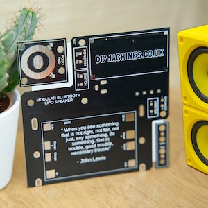 Könnte beinhalten: Eine schwarze Platine mit dem Text "DIYMACHINES.CO.UK" und den Worten "Modularer Bluetooth-LiPo-Lautsprecher". Die Platine enthält ein Zitat von John Lewis: "Wenn du etwas siehst, das nicht richtig, nicht fair, nicht gerecht ist, sag etwas, tu etwas. Gerate in Schwierigkeiten, in gute Schwierigkeiten, in notwendige Schwierigkeiten." Die Platine befindet sich neben einem gelben Lautsprecher mit einem Fragezeichen an der Seite.