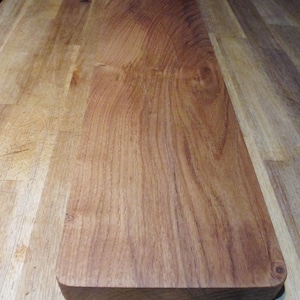 Puede incluir: Una tabla de cortar rectangular de madera con esquinas redondeadas. La tabla tiene un color marrón rojizo intenso con patrones de vetas de madera visibles. La tabla está hecha de una pieza sólida de madera.