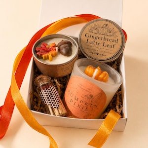 May include: A gift box containing two scented candles and a small glass bottle of matches. One candle is in a brown tin labelled Gingerbread Latte Leaf with a wax maple leaf and acorn. The other is in a glass jar labelled Pumpkin Pixie.