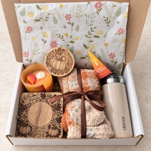 May include: A gift box containing a Pumpkin Magic candle, a round wooden coaster with a pumpkin design, a square wooden coaster with the text hello Autumn, a pair of orange and white fuzzy socks tied with a brown ribbon, and a beige travel mug.