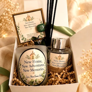 May include: A white gift box containing a frosted glass candle, a glass reed diffuser with black sticks, a green stone, and a card. Both the candle and card feature a house illustration and the text New Home, New Adventures, New Memories.