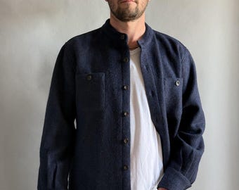 May include: A navy blue shirt jacket with a band collar and button-up front. The jacket has two chest pockets and long sleeves. A white t-shirt is visible underneath. The person is wearing gray jeans.