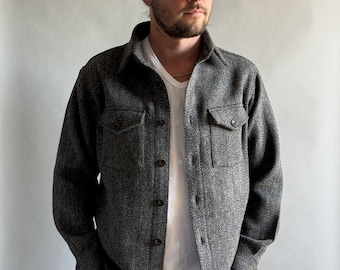 May include: A gray herringbone-patterned shirt jacket with two chest pockets and button closures. The jacket is worn over a white t-shirt and paired with gray jeans. The overall style is casual and classic.
