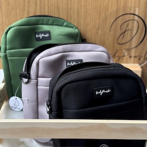 May include: Three small crossbody bags in a wooden crate. The bags are made of a water-resistant material and have a zipper closure. The bags are green, grey, and black. Each bag has a small white logo that says "Daily Paper" on the front.