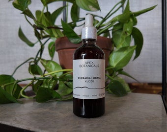 May include: A brown glass bottle with a white label that reads "Apex Botanicals Pueraria Lobata Kudzu Tincture 100ml". The bottle is sitting on a grey surface in front of a green plant.