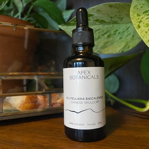 May include: A dark brown glass bottle with a dropper top, labelled "APEX BOTANICALS" and "SCUTELLARIA BAICALENSIS CHINESE SKULLCAP". The bottle is sitting on a wooden surface with a glass box and green plants in the background.
