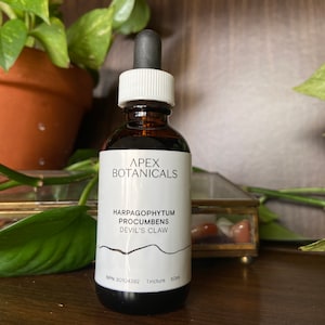 May include: A brown glass bottle with a white dropper top and label. The label reads "Apex Botanicals Harpagophytum Procumbens Devil's Claw" and "NPN 80014392 Tincture 50ml".