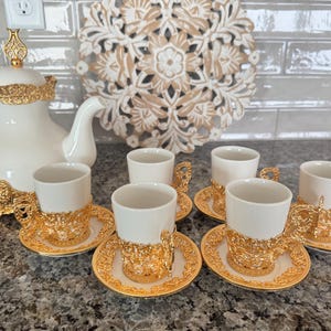 May include: A white porcelain tea set with gold filigree accents. The set includes a teapot, six teacups, and saucers. The teacups and saucers have intricate gold designs. The teapot has a decorative gold handle and lid. The set is designed for serving tea.