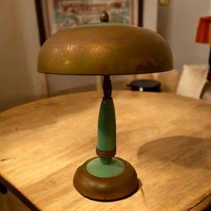 Puede incluir: Una lámpara de mesa vintage verde y dorada con una pantalla en forma de cúpula. La lámpara tiene una base verde con detalles dorados y una pantalla de cúpula dorada.