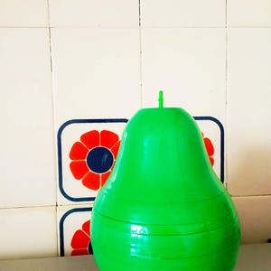 Puede incluir: Un recipiente de plástico de color verde brillante con forma de pera con una pequeña pajita verde que sobresale de la parte superior. El recipiente está colocado sobre una superficie plateada frente a una pared de azulejos blancos con diseños de flores rojas y azules.