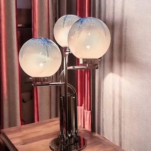Peut inclure: Une lampe de table en chrome avec trois globes en verre blanc, chacun avec un motif en spirale bleu. La lampe est sur une table en bois.