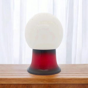 Peut inclure: Une lampe globe blanche avec une base à dégradé rouge et noir. La lampe est posée sur une surface en bois.