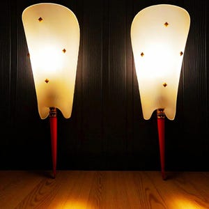 Puede incluir: Dos lámparas de estilo vintage con pantalla crema acanalada y base roja. Las lámparas tienen una forma única y están decoradas con pequeños detalles en forma de diamante. Se encuentran sobre un suelo de madera sobre un fondo oscuro.