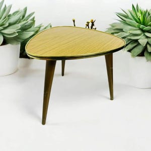 Puede incluir: Una mesa auxiliar de forma triangular, de estilo moderno de mediados de siglo, con una tapa de madera y patas de metal dorado. La mesa está sobre una superficie blanca.
