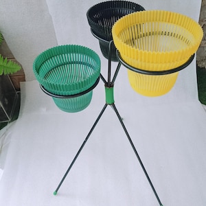 Puede incluir: Un soporte para plantas de tres niveles con estructura de metal negro. Cada nivel sostiene una cesta de plástico acanalada y colorida en verde, amarillo y negro. El soporte tiene una base de trípode con detalles verdes.